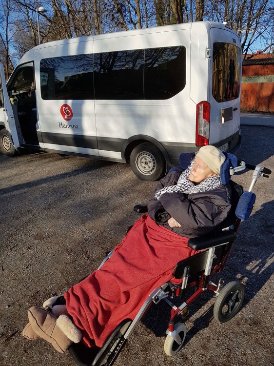 En äldre dam sitter utanför utflyktsbussen i solen